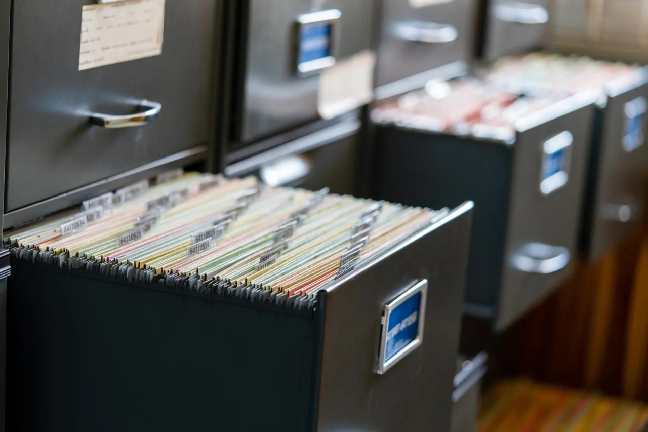 An open filing cabinet drawer of suspension files beside paperwork on a desk