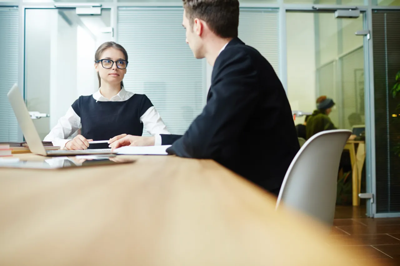 A formal interview across a desk in a glass-walled meeting room