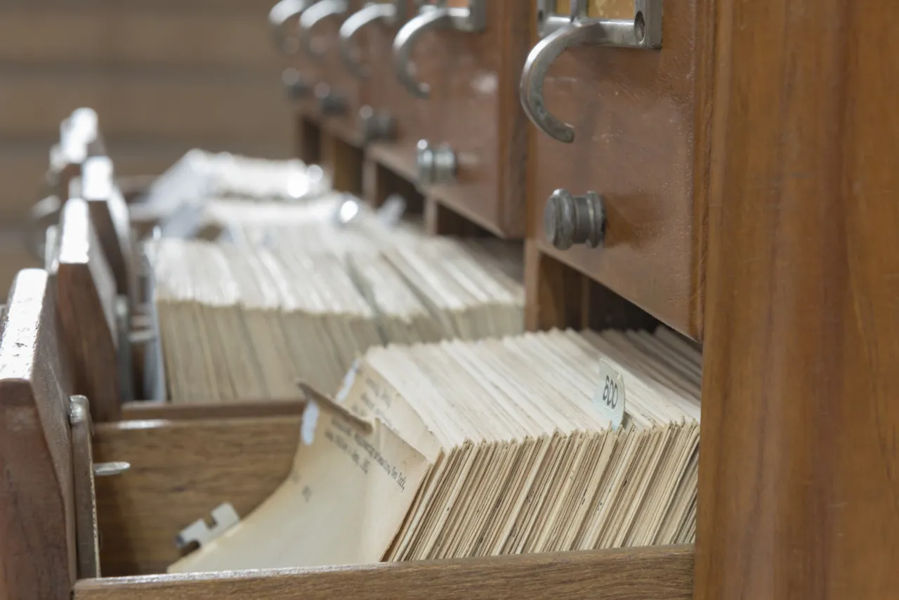 A wooden index card cabinet with one drawer open, holding records