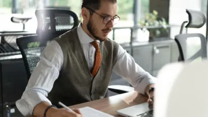 A compliance professional writing notes at a desk in an open-plan office