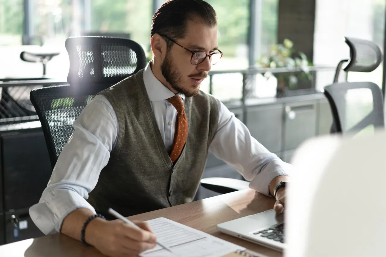 A compliance professional writing notes at a desk in an open-plan office