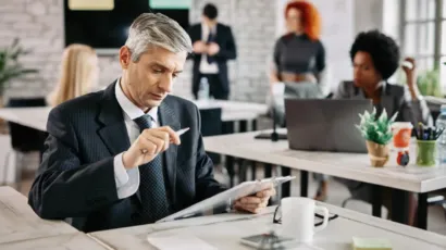 A senior professional reading printed reports at a desk, colleagues working behind