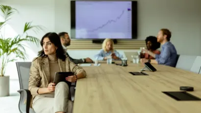 A compliance professional with a tablet at a boardroom table, colleagues seated behind her
