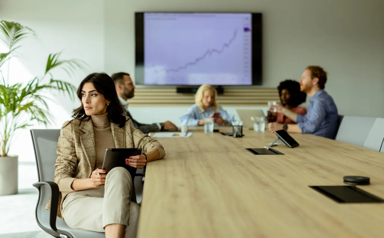 A compliance professional with a tablet at a boardroom table, colleagues seated behind her